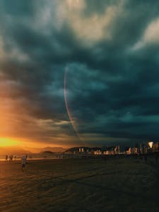 Dramatic sunset on the beach with a visible rainbow and city silhouette under dark clouds.