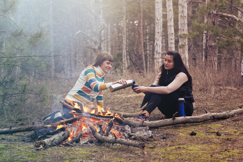 Two women sharing tea and warmth by a bonfire in a serene forest setting.